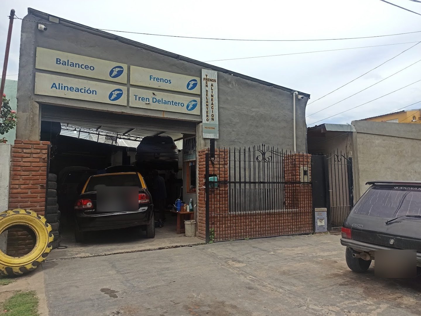 El frente de la gomería sobre la calle, con los carteles de Balanceo, Frenos, Alineación y Tren Delantero, un taxi en el taller y una cubierta amarilla gigante en la vereda