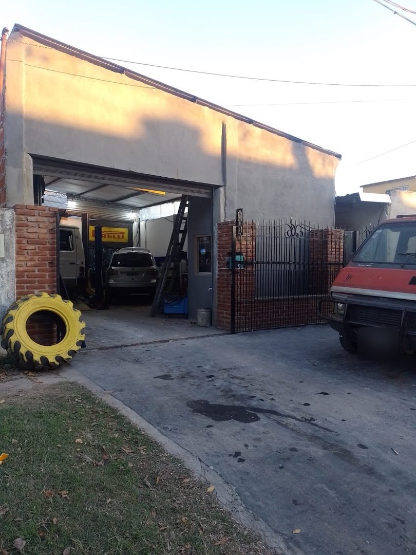 El frente del taller al atardecer: portón negro con ladrillo a la vista, el cartel Pirelli adentro y una cubierta amarilla gigante apoyada en la vereda