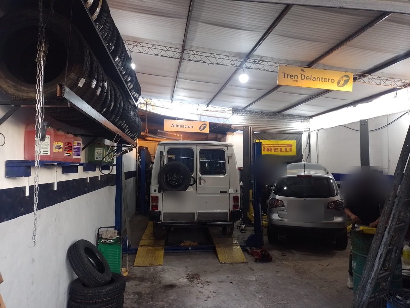 Interior del taller con dos elevadores, un furgón blanco y una camioneta en revisión, cubiertas apiladas en la estantería y los carteles de Alineación y Tren Delantero