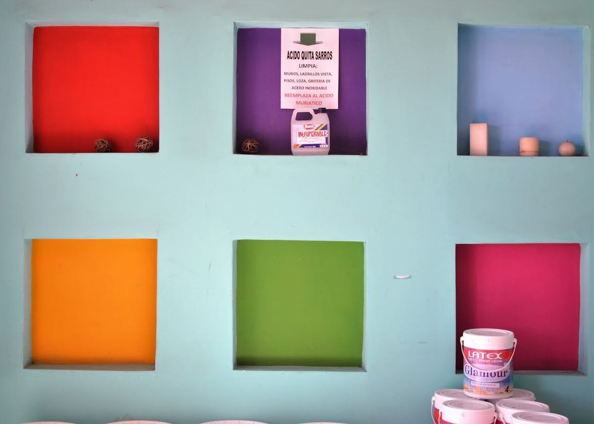 Pared interior de la pinturería con el muestrario de colores pintado a mano, un cartel de ácido quita sarros y baldes de látex Glamour apilados