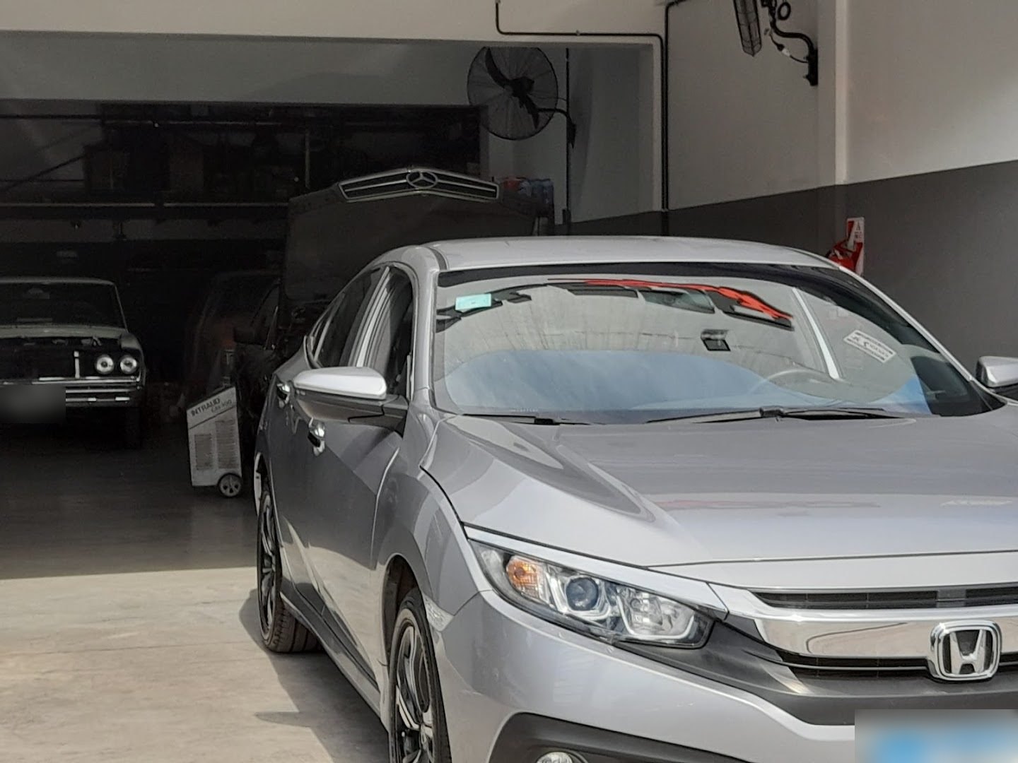 Un Honda Civic gris entra al taller de tu zona; al fondo se ven un auto clásico con el capot abierto y otro con la parrilla de Mercedes