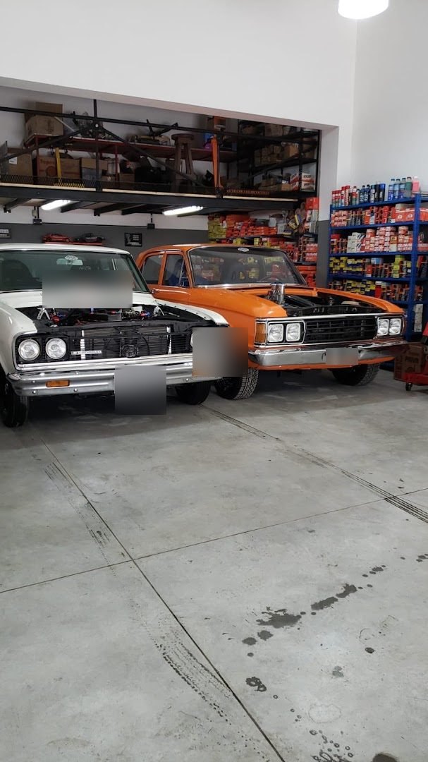 Dos autos clásicos con el capot abierto dentro del taller, uno blanco y otro naranja, con estantes de repuestos al fondo