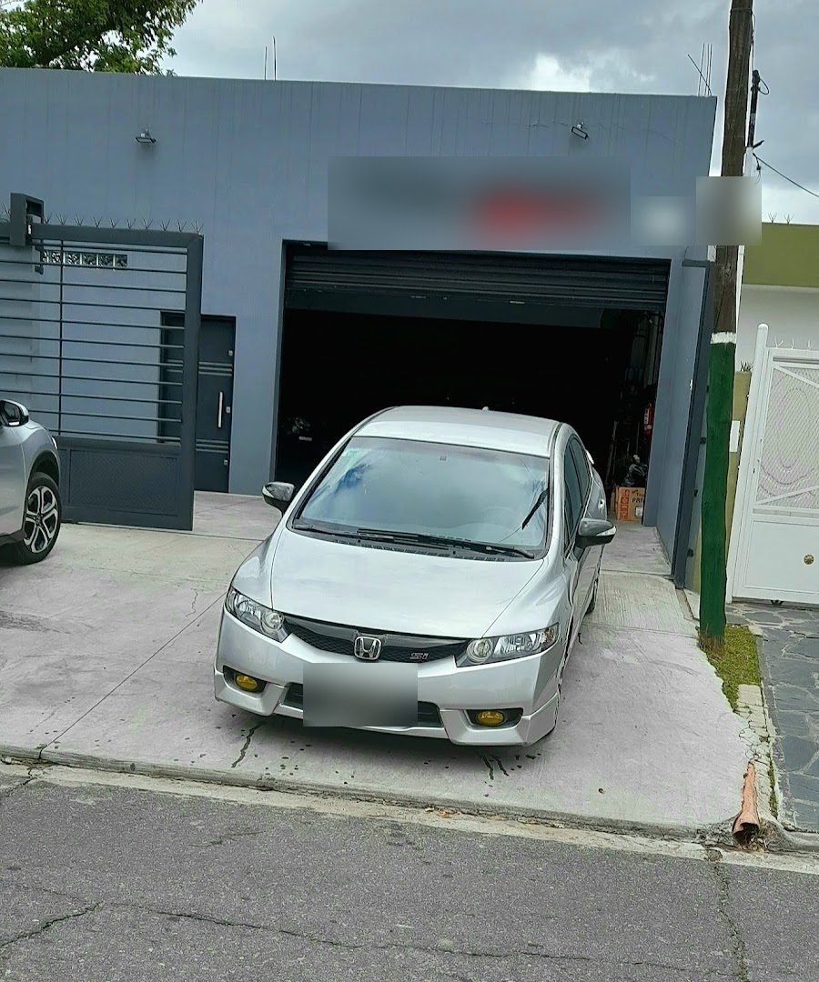 Frente del taller en tu zona: cartel Taller Mecánico sobre la pared gris, portón de chapa y un Honda Civic plateado en la entrada