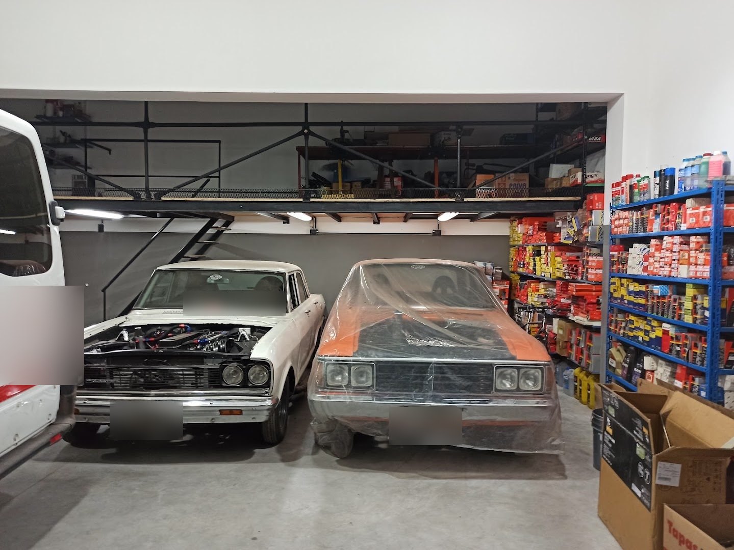 Interior del taller: un clásico blanco con el motor a la vista y otro naranja tapado con nylon, bajo un entrepiso de hierro y con estantes de repuestos a la derecha